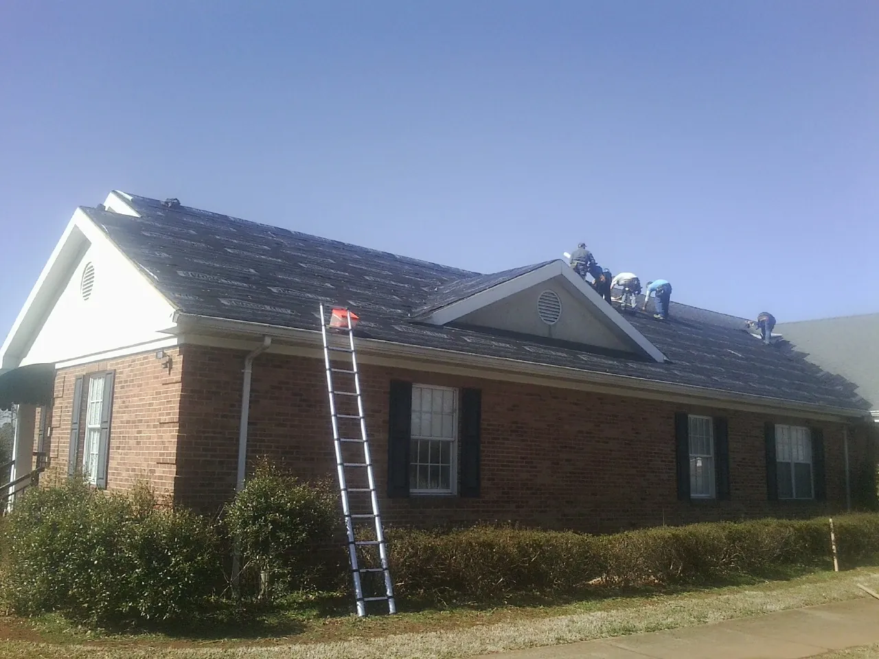 Crew tearing shingles off a brick ranch roof with a ladder set — Metal Roof Repair work by our crew in Harcourt
