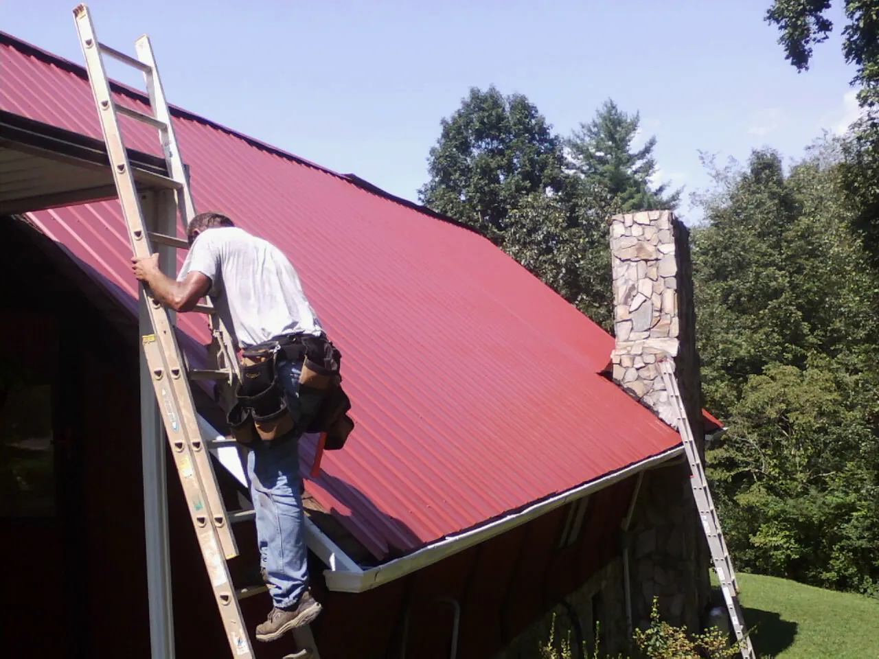Roofer on a ladder beside a red metal roof and stone chimney — Soffit & Fascia Repair by Roofing Brothers in Harcourt